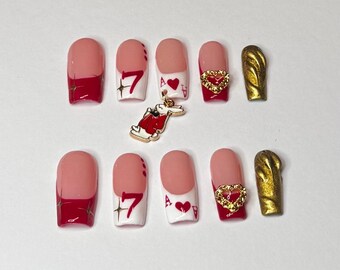 May include: A set of ten artificial nails with a light pink base and red tips. Some nails feature a red "7" design, a heart charm, and a playing card design. One nail is gold. A gold rabbit charm is also present.