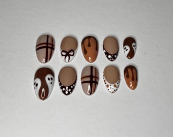 May include: A set of ten oval-shaped false nails with various designs. The nails feature a mix of tan, brown, and white colors. Designs include plaid, bows, dripping paint, polka dots, and ghost-like figures.