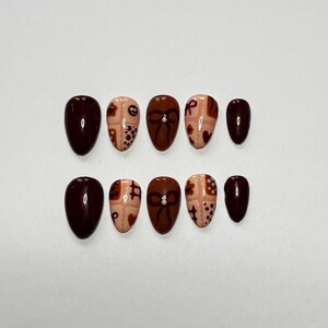 May include: A set of ten oval-shaped false nails. The nails are painted in a variety of designs, including solid dark burgundy, and beige with brown bow and dot patterns. The nails are arranged on a white surface.