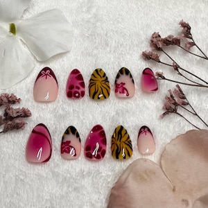 May include: A set of press-on nails with various designs. Some nails feature a pink base with a dark red bow, others have a pink and red pattern, and some have a yellow and black tiger stripe design. The nails are arranged on a white surface with floral accents.