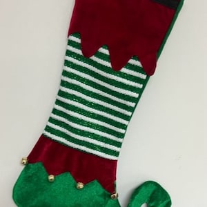 May include: An elf-themed Christmas stocking. It has a red cuff with a gold buckle, a green and white striped body, and a green toe with gold bells. The stocking is made of soft fabric and is designed to resemble an elf's boot.