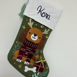Op de afbeelding: Een kerstsok met een groene achtergrond en een witte manchet met de naam "Kora" in marineblauw geborduurd. De sok heeft een cartoonhond met een rode trui, een bot en een ingepakt cadeau.