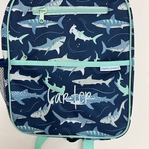 May include: Navy blue backpack featuring a shark design. It has a zipped front pocket and a mesh side pocket. The name "Carter" is embroidered on the front. The brand name "stephen josephe" is also visible.