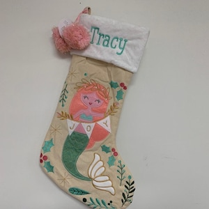 Peut inclure: Une chaussette de Noël beige avec un motif de sirène. La chaussette présente une sirène aux cheveux roses et une queue verte, entourée d'une couronne dorée. Le mot "JOY" est sur une bannière. Le nom "Tracy" est brodé sur le revers blanc, et des pompons roses pendent du haut.