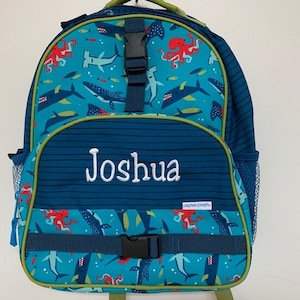 May include: A teal and navy blue backpack with a marine life pattern featuring sharks, whales, and octopuses. The backpack has the name "Joshua" embroidered in white. It includes a front pocket, side mesh pocket, and adjustable straps.