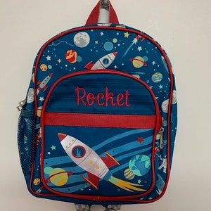 Könnte beinhalten: Ein blauer Rucksack mit Weltraum-Motiv, der Raketen, Planeten und Sterne zeigt. Das Wort "Rocket" ist rot auf der Vordertasche gestickt. Der Rucksack hat rote Verzierungen und eine seitliche Netztasche.