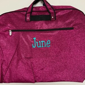 May include: A magenta garment bag with a glittery finish. The bag has a black zip and handles, and the name "June" is embroidered in turquoise. The bag is designed for carrying clothes.