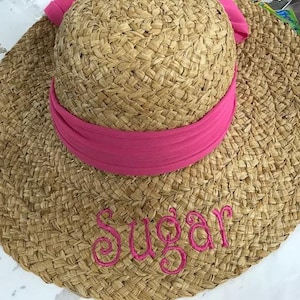 May include: A woven straw sun hat with a wide brim and a pink fabric band around the crown. The word "Sugar" is embroidered in pink script across the brim. The hat is designed for sun protection.