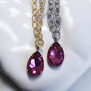 May include: Two necklaces with large, faceted, teardrop-shaped magenta pendants. One necklace has a gold chain, the other a silver chain. The pendants are suspended from a small ring attached to the chain.