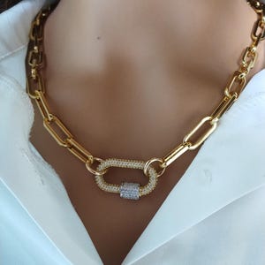 May include: Gold-toned chain necklace with a large, decorative clasp. The clasp is encrusted with small, clear crystals and a central silver-toned bar. The necklace is worn against a white shirt.