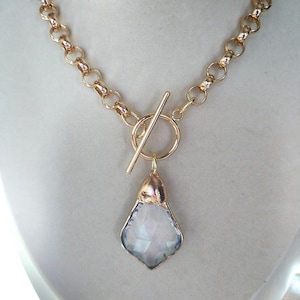 May include: Gold-toned necklace with a large, iridescent pendant. The pendant is a light blue and white gemstone with a gold-colored cap. The necklace features a toggle clasp and a chunky chain.