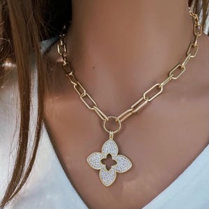 May include: Gold-toned chain necklace with a four-leaf clover pendant. The pendant is encrusted with small, sparkling white stones, and the chain features large, rectangular links. The necklace is worn against a white shirt.