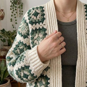 Puede incluir: Un cárdigan de ganchillo con patrón de cuadrados granny en tonos verde oscuro, verde salvia y crema. La prenda tiene una tapeta frontal y puños acanalados en color crema, sobre una camiseta gris oscuro con joyas doradas.
