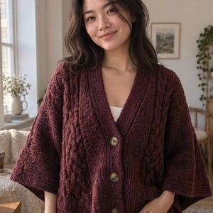 May include: A deep burgundy cable-knit cardigan featuring a V-neckline, wide sleeves, and three large, round, light-colored buttons down the front. The knit texture is prominent throughout the garment, showcasing a classic vertical cable pattern.