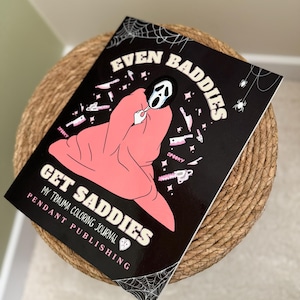 May include: A black and pink colouring book with the title "Even Baddies Get Saddies" and a graphic of a person wearing a ghostface mask and a pink blanket. The book is titled "My Trauma Colouring Journal" and is published by Pendant Publishing.