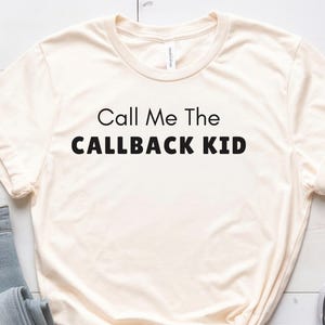 May include: A cream-coloured t-shirt with the text "Call Me The CALLBACK KID" in black. The shirt is styled with blue jeans and navy blue trainers. A cup of coffee and a book are also in the image.