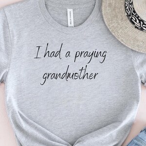 Op de afbeelding: Grijs T-shirt met de tekst "Ik had een biddende oma" in zwarte letters.