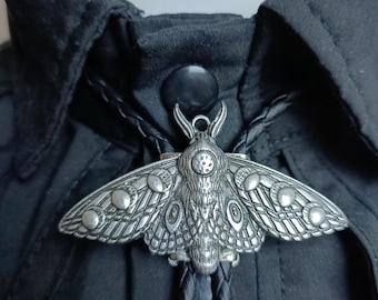 Peut inclure: Une cravate bolo argentée avec un pendentif en forme de papillon de nuit. Le papillon de nuit présente des motifs d'ailes détaillés et un corps texturé. La cravate est faite d'un cordon tressé noir et est présentée sur une chemise noire.