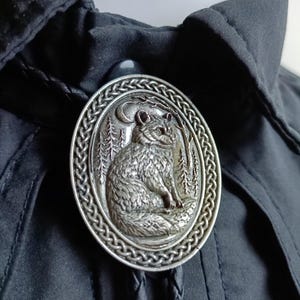 May include: Silver-toned oval bolo tie featuring a detailed relief of a fox seated in a forest under a crescent moon. The fox is surrounded by trees within a braided border. The tie has a black braided cord and a silver-toned clasp.