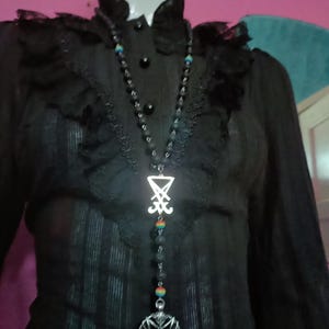 May include: A long necklace featuring black textured beads, small rainbow-coloured beads, a silver-toned Sigil of Lucifer pendant, and a circular silver-toned pendant depicting a goat head within a pentagram, worn over a black ruffled blouse.