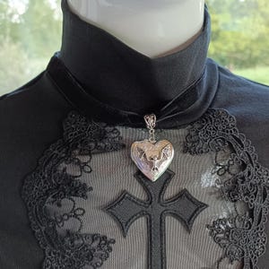 Peut inclure: Un collier pendentif argenté en forme de cœur avec un motif de chauve-souris, suspendu à un ras du cou en velours noir. Le collier est présenté sur un haut noir avec des détails en dentelle et une croix.