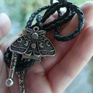 May include: A silver-toned bolo tie featuring a moth pendant with crescent moon details. The pendant is attached to a black braided cord with decorative silver tips and ball ends. The bolo tie is a unique accessory.