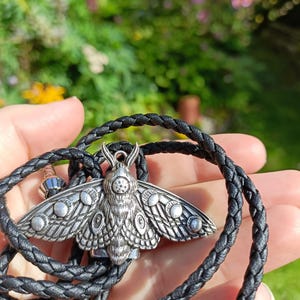 May include: A silver moth pendant on a black braided cord. The moth pendant has detailed wings and body. The cord is thick and appears to be leather. The background is blurred, with green foliage and sunlight.