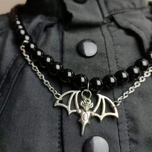 Op de afbeelding: Een zwarte kralenketting met een zilveren vleermuis hanger. De ketting heeft een dubbele rij zwarte kralen en een zilveren ketting. De vleermuis hanger is gedetailleerd met uitgespreide vleugels en een centraal lichaam. De ketting wordt getoond op zwarte stof.