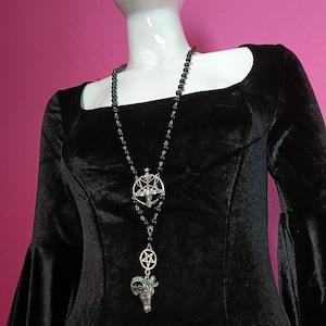 Peut inclure: Un long collier composé de perles noires et de breloques en métal argenté. Le design comprend un emblème pentagramme circulaire et un pendentif en forme de tête de bélier suspendu à une chaîne. Le collier est présenté sur un haut noir en velours.