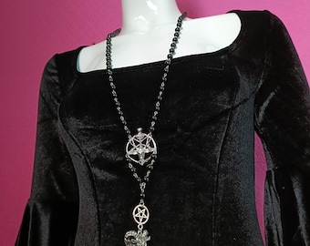 May include: A long necklace featuring black beads and silver-toned metal charms. The design includes a circular pentagram emblem and a detailed ram head pendant suspended from a chain. The necklace is displayed on a black velvet long-sleeve top.