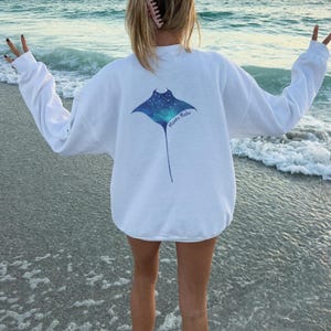 May include: White sweatshirt with a blue and green manta ray graphic on the back. The graphic has a starry sky design and the text "Manta Babe" underneath.