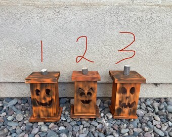 May include: Three wooden jack-o'-lantern candle holders with carved faces. The candle holders are painted in shades of brown and orange.