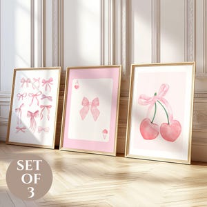 Puede incluir: Conjunto de tres láminas artísticas enmarcadas. Las láminas presentan ilustraciones en acuarela de lazos rosas, una carta de juego con un lazo y cerezas con un lazo. Los marcos son dorados y el fondo es rosa claro.