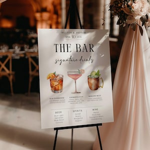 以下が含まれることがあります： カクテルのイラストと黒い文字が書かれた白い看板。看板には「The Bar Signature Drinks」というタイトルが付けられており、「オールドファッションド」「コスモポリタン」「モスクワミュール」というドリンクがリストされています。看板にはビール、スピリッツ、ワインの選択肢も記載されています。
