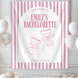 Puede incluir: Un tel&oacute;n de fondo a rayas rosas y blancas con el texto "WELCOME TO EMILY'S BACHELORETTE" en escritura rosa. Dos copas de champ&aacute;n con lazos aparecen debajo del texto, con "MIAMI, FLORIDA" en la parte inferior. Globos blancos en primer plano.