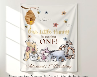 May include: A white banner featuring Winnie the Pooh and friends, with a beehive, bees, and flowers. Text reads "Our Little Hunny is turning ONE!" and "Adriana's 1st Birthday." Customizable with name and age, available in multiple sizes.