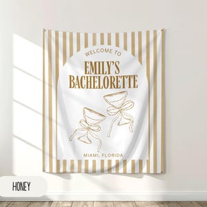 Puede incluir: Un tapiz rectangular con fondo blanco y rayas verticales beige. El centro presenta el texto "Welcome to Emily's Bachelorette" con dos copas de champ&aacute;n y la ubicaci&oacute;n "Miami, Florida". La palabra "HONEY" est&aacute; en un rect&aacute;ngulo blanco en la parte inferior.