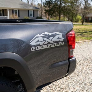 Op de afbeelding: Donkergrijze truckbed met een witte 4x4 OFF ROAD sticker. De sticker heeft een bergketenontwerp boven de tekst. Het achterlicht van de truck is zichtbaar aan de rechterkant. De truck staat op een grindoprit.