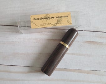 May include: A dark brown wooden needle case with a gold-colored band. The case is cylindrical and has a smooth finish. The text "Needlework Accessories Schandworks" is visible on a small label.
