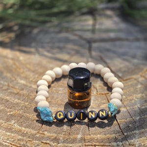 Puede incluir: Una pulsera de cuentas de madera con el nombre "QUINN" escrito con cuentas de letras negras y doradas. La pulsera presenta detalles de piedra turquesa y una pequeña botella de aceite esencial de vidrio ámbar con tapa negra.