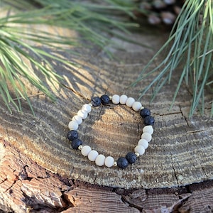 May include: A beaded bracelet featuring alternating white and black lava stone beads, interspersed with small gold-colored beads. The bracelet is displayed on a rustic wooden surface, with pine needles and cones in the background.