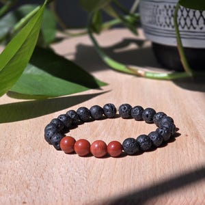 Könnte beinhalten: Ein Armband mit schwarzen Lavasteinperlen und roten Jaspisperlen. Das Armband wird auf einer hellbraunen Holzoberfläche präsentiert. Der Hintergrund umfasst grüne Blätter und einen dekorativen Topf.