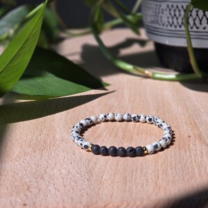 May include: A beaded bracelet featuring white and black spotted beads, black lava beads, and gold-colored accents. The bracelet is displayed on a wooden surface with green leaves and a decorative pot in the background.