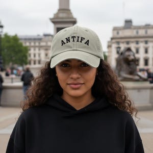 Peut inclure: Casquette de baseball vert sauge avec le mot "ANTIFa" brodé en lettres noires. Le chapeau est porté par une personne portant un sweat à capuche noir. L'arrière-plan présente un ciel nuageux et un bâtiment.