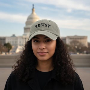 Op de afbeelding: Lichtgroene baseballcap met het woord "RESIST" in zwart geborduurd. De pet wordt gedragen door een persoon voor een gebouw met een koepel. De persoon draagt een zwarte jas.