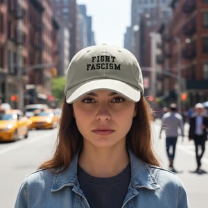 Op de afbeelding: Lichtgroene baseballcap met de tekst "FIGHT FASCISM" in zwarte letters. De cap wordt gedragen door een persoon in een spijkerjasje, met een stadsstraat op de achtergrond.