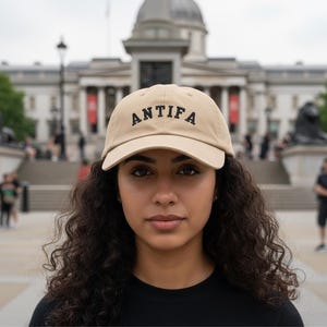 Op de afbeelding: Beige baseballpet met het woord "ANTIFIA" in zwart geborduurd. De pet wordt gedragen door een persoon voor een gebouw met zuilen en een grote koepel. De persoon draagt een zwart shirt.