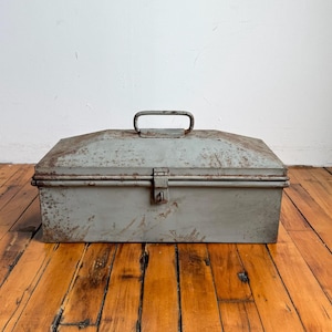 May include: A weathered, rectangular metal toolbox in a muted gray hue. The box features a hinged lid with a handle and a latch closure. The metal shows signs of rust, adding to its vintage aesthetic. The toolbox sits on a wooden surface.