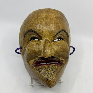 May include: A carved wooden mask with a light brown finish, featuring a pointed nose, arched black eyebrows, and a mouth painted with dark red lips. The mask has a textured surface and includes purple elastic cords attached to the sides for securing.