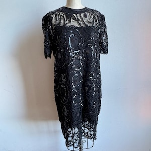 May include: Black vintage dress with short sleeves, intricate lace detailing, and sequin embellishments. The dress features a round neckline and a knee-length hemline. The lace pattern includes circular and floral designs, creating a textured appearance. The dress is displayed on a mannequin.